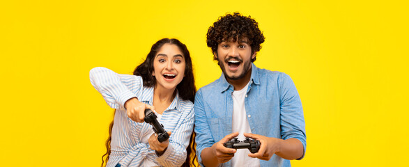 Two friends are having fun playing video games in a yellow room. They are holding game controllers and smiling at the camera. Their expressions show excitement and joy. © Prostock-studio