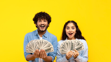 A young couple stands together holding fan-like stacks of cash. They display excited expressions while posing against a bright yellow background. They are dressed casually. © Prostock-studio