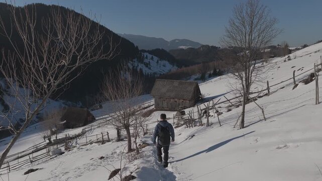 Cinematic tracking shot of a happy hiker walking down a snowy mountain trail among rustic barns at sunset, ungraded log