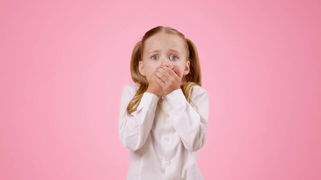 A young girl with light skin and blonde hair stands against a pink background. She appears shocked and surprised, with wide eyes and an open mouth. Her hands are clasped in front of her.