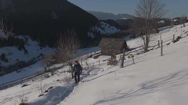 Cinematic right panning shot of a hiker walking on a snowy mountain trail among rustic barns at sunset, ungraded log
