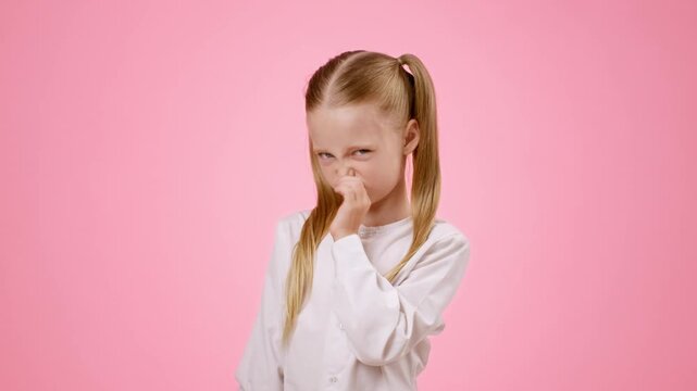 A young girl with light skin and long blonde hair stands in front of a pink background. She has a playful pout and is making a funny face with her hand raised as if signaling to stop.