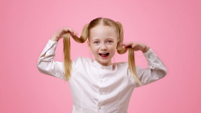 A girl with blonde hair styled in pigtails is having fun pulling her hair while smiling. She wears a white long-sleeve shirt and is posed against a pink background, showcasing joy and playfulness.