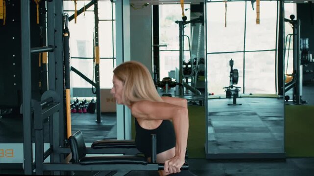 A woman with light skin is doing dips on gym equipment. She focuses on her exercise in a bright and spacious gym setting. Mirrors reflect her movements while various machines are visible.