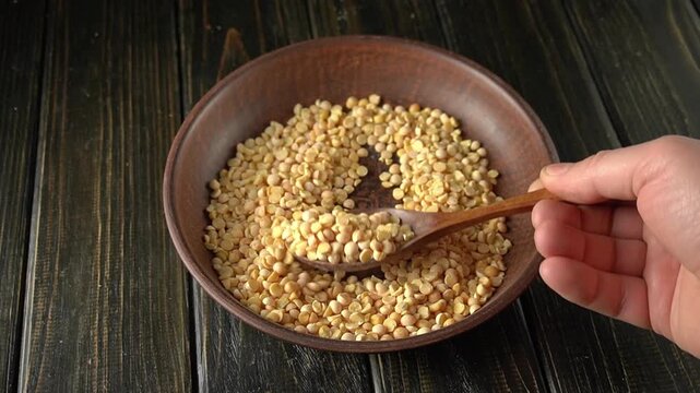 Hand scooping dried yellow split peas from a wooden bowl with a wooden spoon, showcasing the gradual movement of the pea across a rustic wooden surface