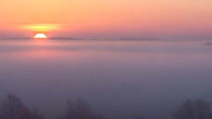 Fototapeta premium The sun rising over a dense sea of fog in a rural valley, vibrant orange sun peeking through misty horizon, atmospheric autumn morning scenery