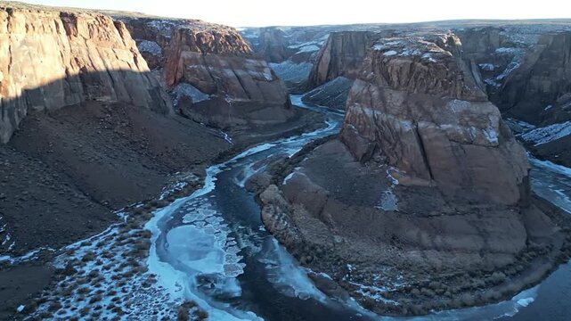 Dramatic aerial view of a winding river cutting through a majestic desert canyon, with icy patches along the banks, suggesting a cold season.