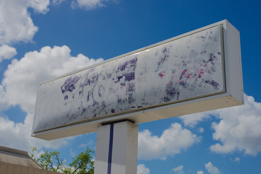 A faded outdoor billboard sign mounted on a pole features a blank, weathered advertising panel with peeling paint and ghosted graphics against a bright blue sky with scattered clouds.
