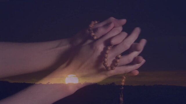 Holding rosary beads, hands silhouetted against sunset in peaceful landscape