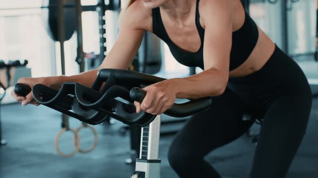 A woman with light skin is engaged in a workout on a stationary bike inside a bright gym. She focuses on maintaining her form as she pedals, surrounded by gym equipment.