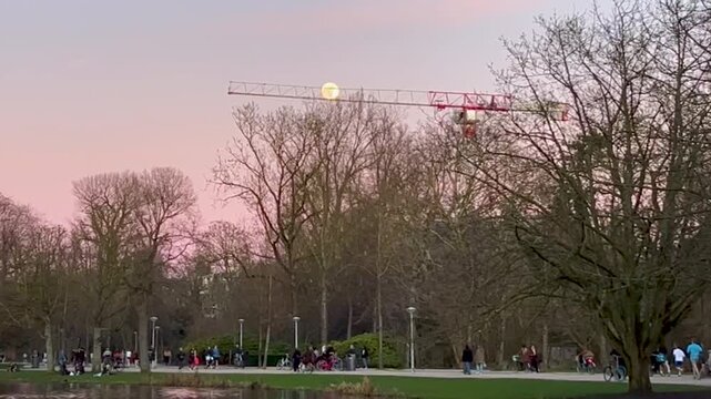 People Exercising in City Park at Sunset with Full Moon and Construction Crane