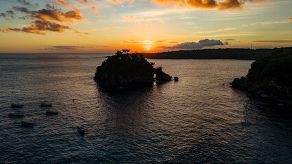 Drone shot of small rocky island with natural arch surrounded by boats at sunset