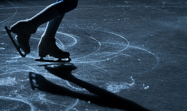 Close up of figure skater legs and blades on a shiny indoor ice rink with reflection and shadow. Elegant winter sport scene showing balance, motion and training.