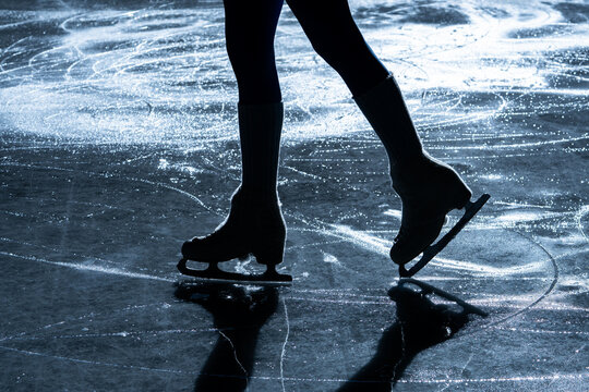 Close up of figure skater legs and blades on a shiny indoor ice rink with reflection and shadow. Elegant winter sport scene showing balance, motion and training.