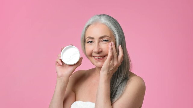 A woman with gray hair smiles as she applies cream to her face. She holds a jar of skincare product in her other hand. The background is a soft pink color, creating a bright setting for her routine.