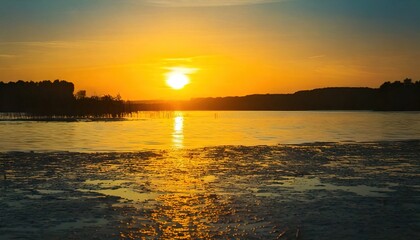 Obraz premium A vibrant sunset over a calm lake, with trees silhouetted against the bright horizon