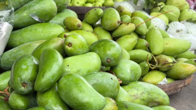Large unripe mangoes lie tightly packed across a display surface, with surrounding stalls and customer movement shaping a busy retail setting. Fresh mangoes.