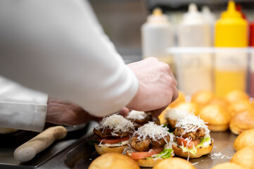 A chef meticulously finishes several gourmet sliders on a kitchen prep station, garnishing them...
