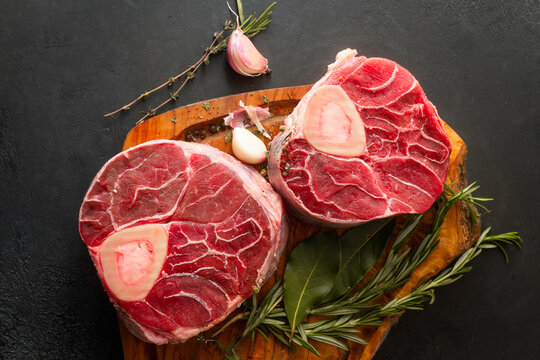 Raw osso buco cuts with bone, placed on a rustic wooden cutting board, accompanied by fresh rosemary, thyme, and garlic cloves. Top view.