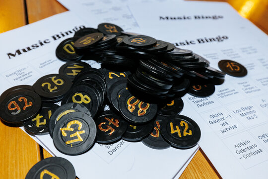 Black bingo chips with numbers scattered on a wooden table alongside printed music bingo cards featuring song titles and artists