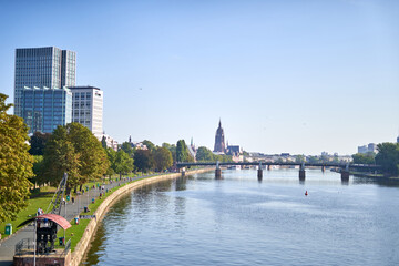 Obraz premium A scenic panoramic view of Frankfurt am Main, Germany, showcasing the Main River flowing through the city center on a bright sunny day. The left riverbank features a pedestrian promenade lined with