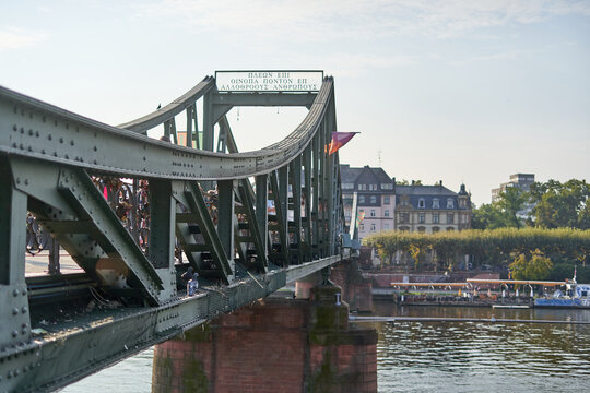 The image captures the iconic Eiserner Steg (Iron Bridge) in Frankfurt am Main, Germany. This historic steel truss pedestrian bridge spans the River Main, featuring its characteristic greenish-gray p