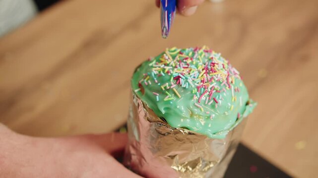 Male hand gently adding colorful sprinkles to green frosted easter cupcake from bag. Closeup of foilwrapped base on wooden table. Festive party vibe with decorations in home kitchen