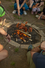 Obraz premium Group grilling sausages around firepit at twilight, parents and kids tending skewers, orange flame flicker, picnic style seating. People celebrate the traditional Czech holiday Carodejnice outdoors.