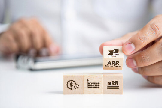Business professional arranging wooden blocks with financial symbols and terms like MRR and ARR for growth analysis