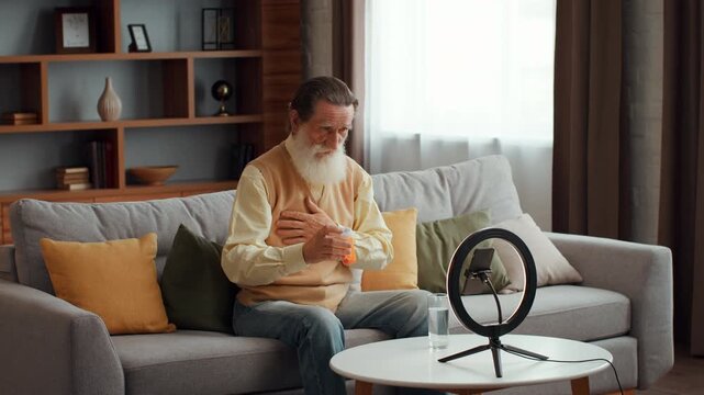 Senior bearded man recording video instruction about first medical aid if having heart attack, touching chest and showing pills to smartphone camera, slow motion, free space