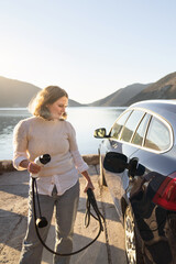 Fototapeta premium Woman traveler with charging plug next to her electric car on the sea coast at sunset