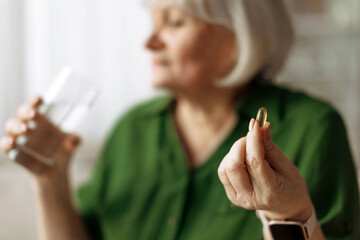 Senior woman holding pill and glass of water while sitting at home. Mature caucasian female taking...