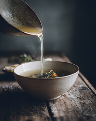 Clear vegetable soup poured into ceramic bowl with herb garnish creating warm steam and rustic mood