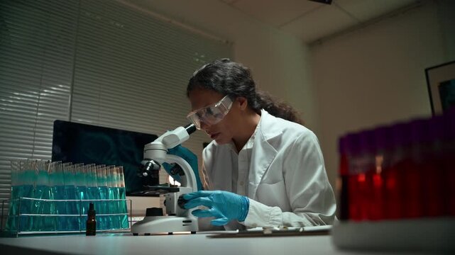 Focused female scientist in protective glasses working on a scientific experiment. A microbiologist using a microscope in a dark laboratory