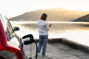 Fototapeta premium Woman with smartphone next to charging electric car on the seacost during summer vacation