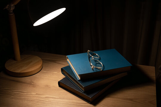 Stack of books and eyeglasses under warm lamp light in dark room.