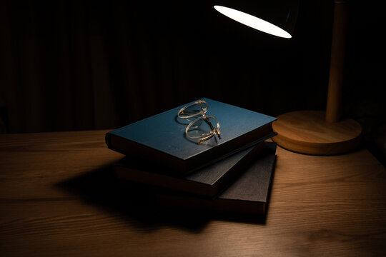 Stack of books and eyeglasses under warm lamp light in dark room.