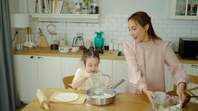 Happy Asian mother and cute little daughter baking together in a modern kitchen. Woman pouring flour into a sifter held by the child. Family bonding, cooking at home, and learning concept.