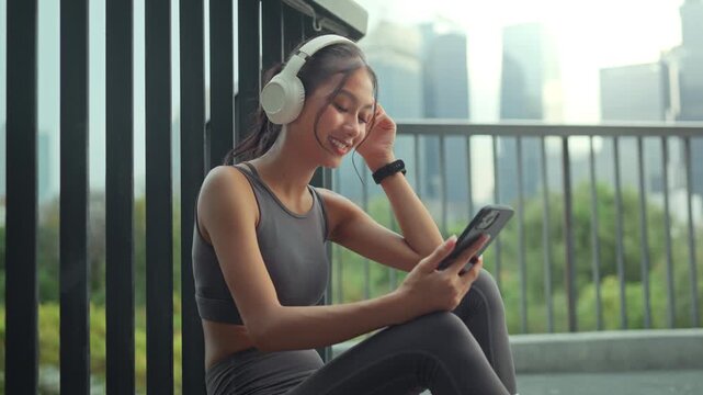 Happy Asian woman in fitness apparel with white headphones using smartphone in urban park. Young girl relaxing with music and wellness app on balcony, city lifestyle, workout break, and personal relax