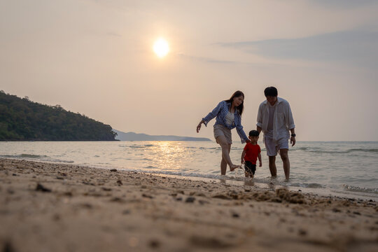 Happy Asian family with a young son enjoying sunset on the beach, playing in the sand and walking together during summer vacation.