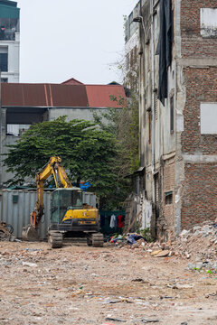 Demolition site in Hanoi, Vietnam: yellow excavator amidst rubble and debris, surrounded by weathered brick buildings with exposed walls and hanging laundry, showcasing the juxtaposition of urban deca