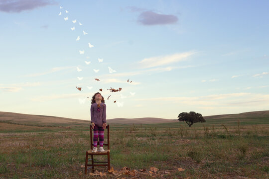 Girl standing on a chair in the countryside looking up at autumn leaves mid air turning into butterflies