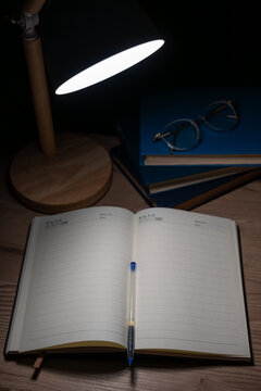 Open blank notebook with pen on desk near stack of books under lamp light.