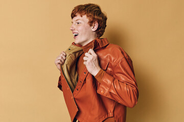 Young man with red hair wearing orange leather jacket smiles and poses with closed eyes on beige...