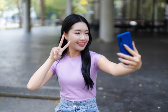 Young Asian woman smiles taking selfie with smartphone making peace sign gesture outdoor public space for social media casual modern lifestyle