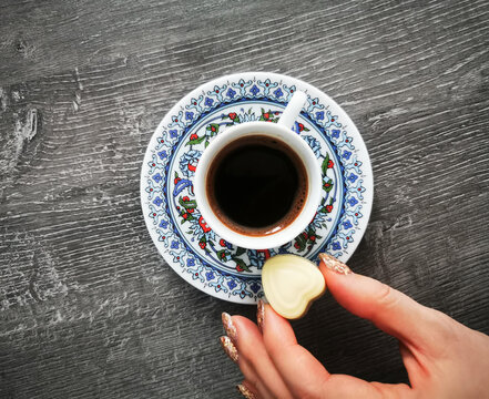 A decorative cup of coffee sits on a patterned saucer as a hand places a chocolate heart beside it. This image represents warm drinks, sweet details, and relaxed coffee rituals.