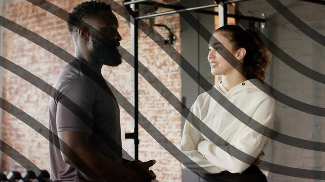 Trainer gesturing and explaining by rig in fitness, client uncrossing arms, leaning closer for tips