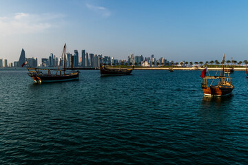 Fototapeta premium Wooden dhows float on the calm waters of Doha, Qatar, with the city’s iconic modern skyline highlighting the contrast between heritage and contemporary urban life. 