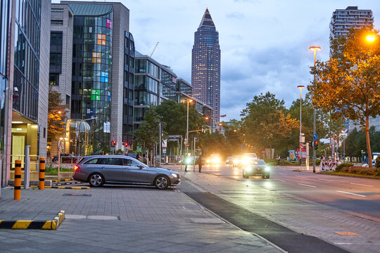 Urban evening scene in Frankfurt am Main, Germany, featuring the iconic Messeturm (Trade Fair Tower) skyscraper rising prominently in the background against a cloudy twilight sky. Modern glass and st