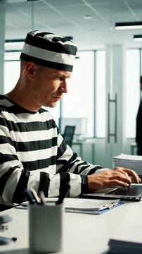 Prisoner working on laptop in modern office. He focuses on data entry while surrounded by paperwork. Vertical shot captures determination and productivity.